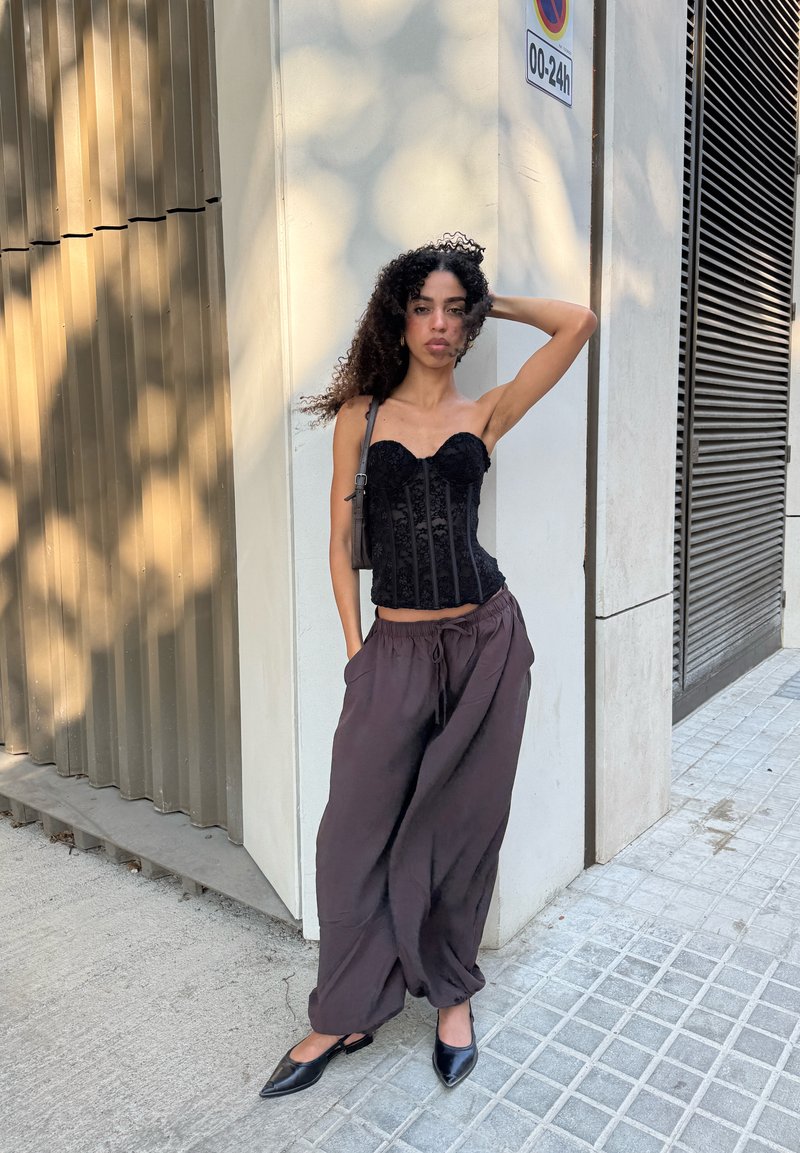 Femme aux cheveux bouclés portant un haut corset noir et un pantalon ample foncé, appuyée contre un coin de bâtiment sur un trottoir carrelé.