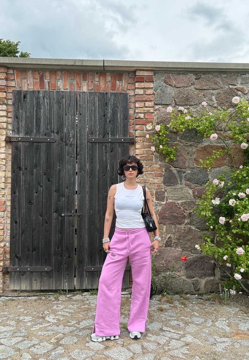 Personne aux cheveux noirs et lunettes de soleil portant un débardeur blanc et un pantalon rose se tient sur un chemin de pierres près d'une porte en bois et d'un mur en briques avec un buisson de roses en fleurs.
