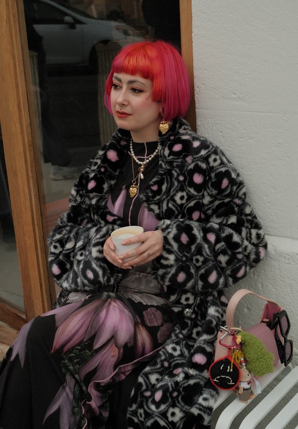 Persoon met felroze bobkapsel en patroonmantel zittend, met een kopje in de hand, draagt bloemige broek en laagjes sieraden, met een roze tas in de buurt.