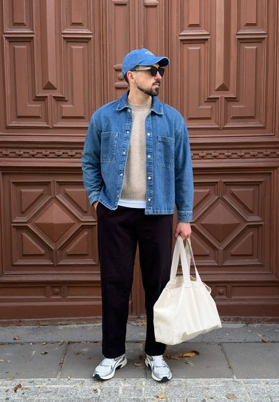 Chaqueta de mezclilla sobre un suéter beige, pantalones negros, zapatillas gris y blancas, y llevando una bolsa de tote de color claro frente a un fondo marrón texturizado.