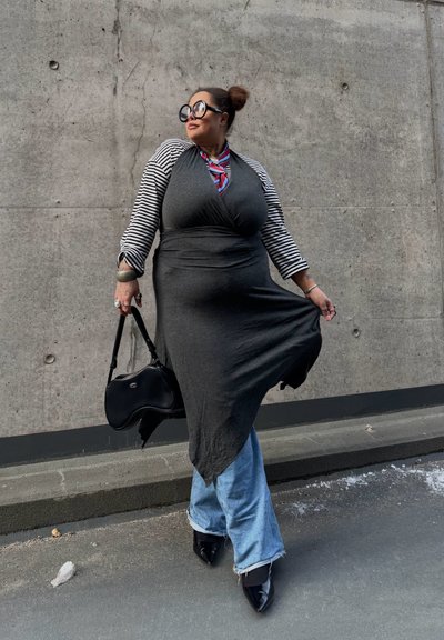 Mujer con grandes gafas redondas, mangas a rayas, vestido largo oscuro tipo túnica sobre jeans, sosteniendo una bolsa negra, posando frente a una pared de concreto.