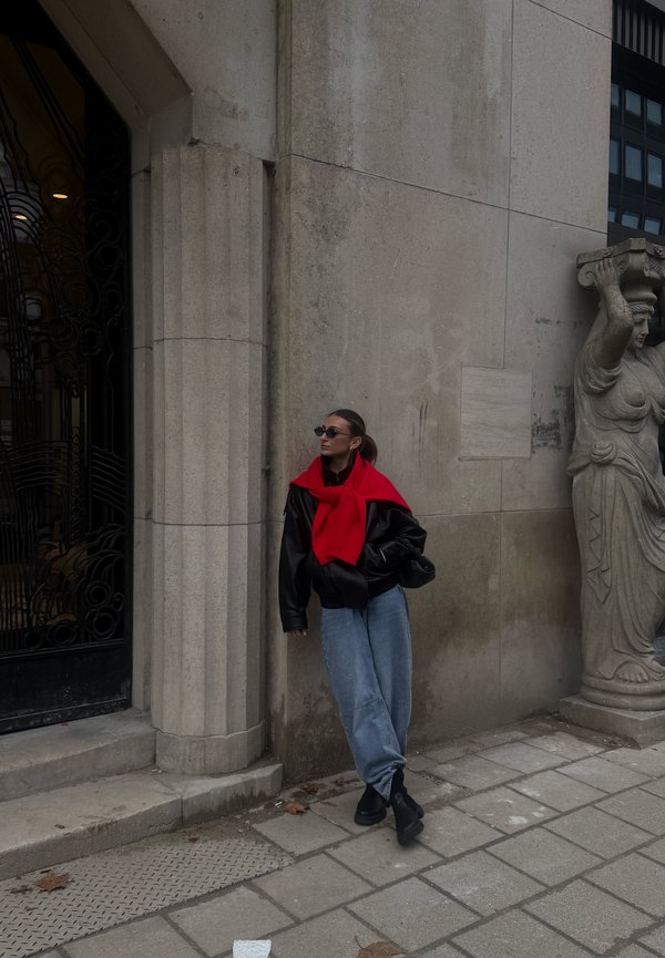 Une femme portant des lunettes de soleil, un pull rouge, une veste noire et un jean ample s’appuie contre un bâtiment en pierre, à côté d’une porte ornée et d’une statue sculptée.