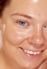 Närbild på ett ansikte med slät, något glansig hud som har fräknar. Framträdande blå ögon, näsring och ett lätt leende med synliga tänder.