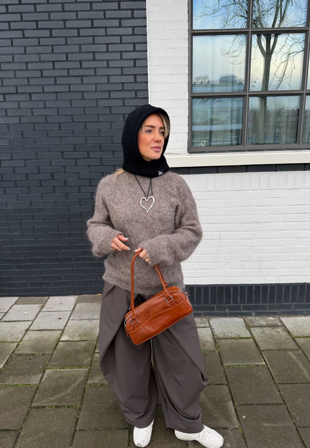 Femme portant une capuche noire, un pull brun pelucheux, un pantalon large gris, des baskets blanches, un collier en forme de cœur, tenant un sac à main marron, debout sur un trottoir devant un mur en briques noir et blanc.