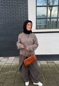 Femme portant une capuche noire, un pull brun pelucheux, un pantalon large gris, des baskets blanches, un collier en forme de cœur, tenant un sac à main marron, debout sur un trottoir devant un mur en briques noir et blanc.