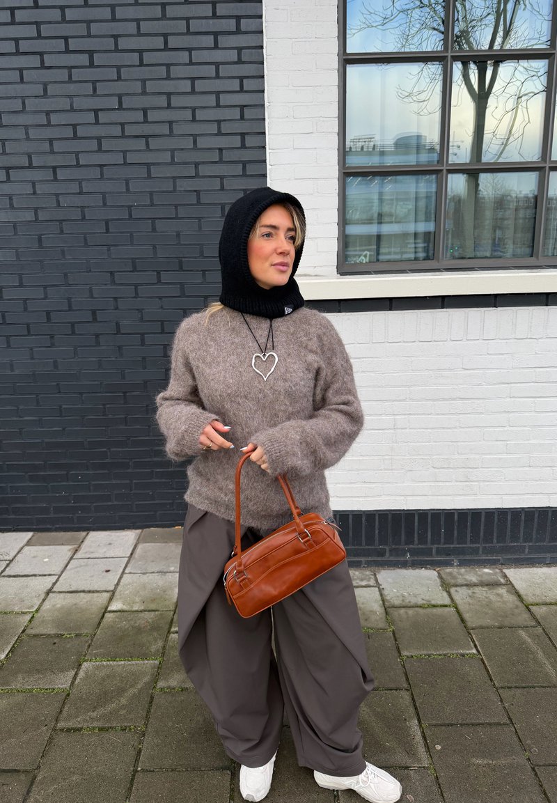 Femme portant une capuche noire, un pull marron pelucheux, un pantalon large gris, des baskets blanches, un collier en forme de cœur, tenant un sac à main marron, debout sur le trottoir près d'un mur en briques noir et blanc.