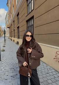 Mörkbrun ulljacka med vida ärmar och knappdetaljer, kombinerad med en brun polotröja. Bär en liten brun mockaväska.