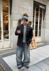 Black oversized blazer, grey sweater, and wide blue denim jeans. Holding a coffee cup while standing on a pavement near a shop adorned with plants.
