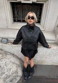 Schwarze übergroße Lederjacke mit hohem Kragen, kombiniert mit einem mehrlagigen schwarzen Rock und spitzen schwarzen Stiefeln. Kleine metallische Handtasche auf einem Steinsims.