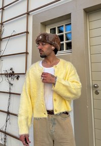 Άνδρας με γυαλιά, γούνινο καπέλο, κίτρινη ζακέτα και μπεζ παντελόνι στέκεται κοντά σε έναν τοίχο με παράθυρο και πόρτα, κοιτάζοντας προς τα πλάγια.