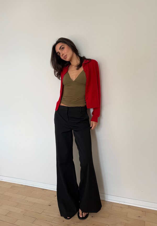 Mujer de cabello oscuro apoyada en la pared, vistiendo un cárdigan rojo, camiseta sin mangas verde oliva, pantalones negros de pierna ancha y chanclas negras sobre suelo de madera.