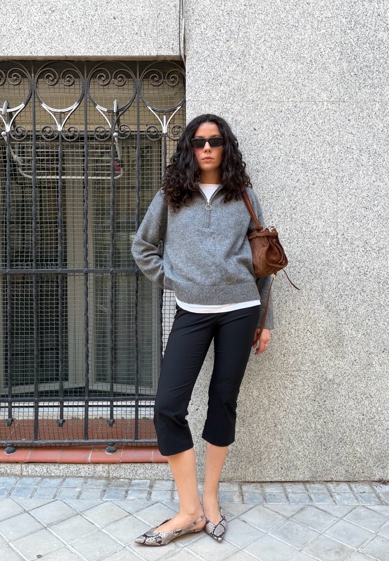 Donna con capelli ricci, che indossa occhiali da sole, un maglione grigio, pantaloni neri a vita alta, ballerine in pelle di serpente e una borsa a tracolla marrone, in piedi contro un muro di pietra.