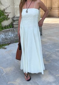 Weißes, ärmelloses Kleid mit tailliertem Oberteil und schwingendem Rock, aus leichtem Stoff gefertigt. Eine geflochtene braune Tasche und schwarze Flip-Flops runden den Look ab.