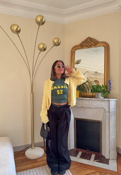 Cardigan de punto amarillo, top corto verde con texto "BRASIL 92", pantalones plisados negros, sosteniendo un bolso negro; interior moderno con lámpara dorada y espejo.