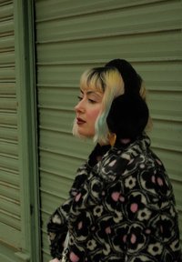 Svart og rosa blomster-mønstret faux fur frakk med høy krage, parret med svarte øreklokker og en sølvkjedeveske, mot en grønn bakgrunn.