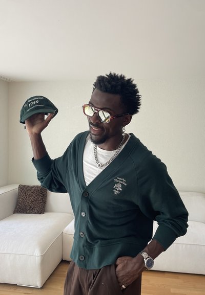 Hombre con cárdigan oscuro y gafas de sol sostiene una gorra verde con texto blanco, de pie en una sala de estar con un sofá blanco y un cojín con estampado de leopardo.