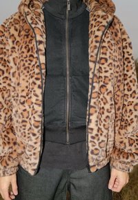 Veste en fausse fourrure à imprimé léopard avec capuche, couche intérieure zippée noire contrastante, poignets côtelés et coupe décontractée, associée à un pantalon foncé.