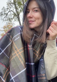 Poncho a quadri in tonalità verde scuro, nero e marrone con cappuccio. Il materiale sembra leggero e impermeabile, con un logo sul lato.
