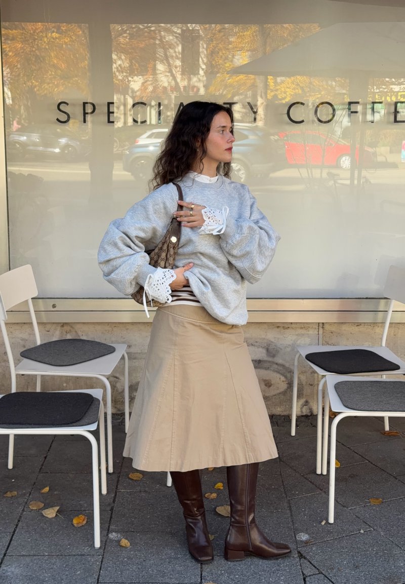 Sudadera gris claro con mangas abullonadas, puños de encaje blanco, falda beige en línea A y botas de cuero marrón hasta la rodilla, ambientado en una cafetería al aire libre.