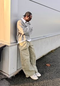 Grå genser lagt over en hvit skjorte, løse lysegrønne cargobukser og hvite joggesko. Flettet frisyre mot en grå vegg.