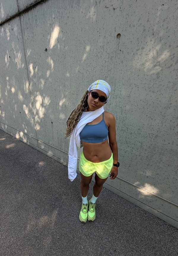 Frau in Sportkleidung mit neongelben Shorts und Schuhen, blauem Sport-BH, weißem Kopftuch und Handtuch steht draußen an einer Betonwand.