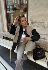 Giacca di pelle nera con colletto in maglia beige, sneakers grigie, pantaloni beige chiaro e borsa scura con texture e dettagli dorati.