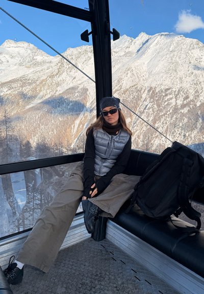 Joven mujer con gafas de sol y ropa de invierno sentada dentro de un teleférico con montañas nevadas visibles fuera de la ventana.