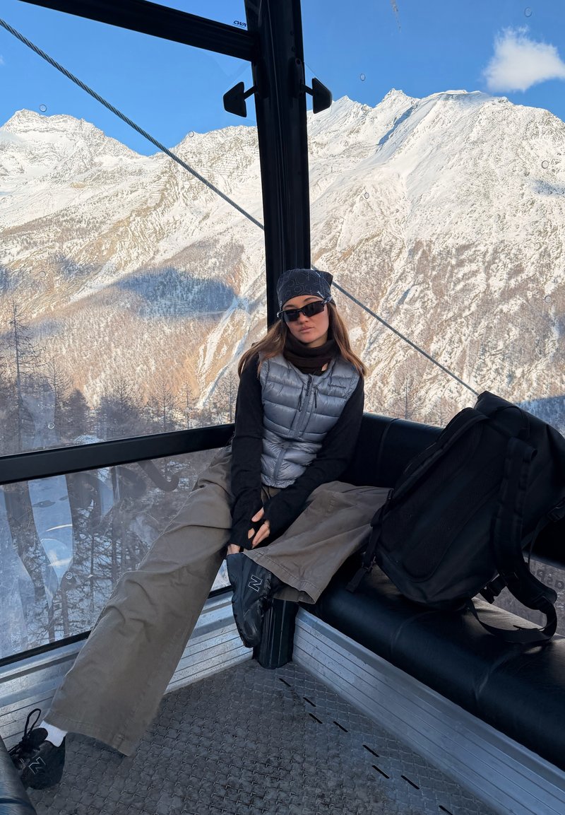 Jeune femme portant des lunettes de soleil et des vêtements d'hiver, assise à l'intérieur d'une cabine de téléphérique avec des montagnes enneigées visibles à travers la fenêtre.