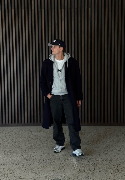 Abrigo de marinero, sudadera gris, camiseta blanca con logotipo, vaqueros negros y zapatillas blancas. La persona está apoyada contra una pared de madera con tablones verticales.