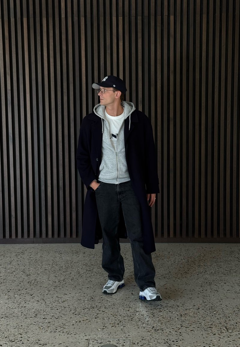 Marineblauwe overjas, grijze hoodie, wit T-shirt met logo, zwarte spijkerbroek en witte sneakers. Persoon staat tegen een houten muur met verticale latten.