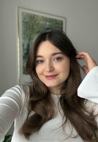 Portrett av en person med langt, bølget brunt hår, iført en hvit ribbestrikket genser med lange ermer, smilende; myk belysning og en naturinspirert bakgrunn synlig.