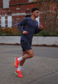 Tenue de sport pour homme comprenant une chemise à manches longues bleu marine, un short bleu marine et des chaussures de course orange fluo avec des accents blancs et des semelles vertes.