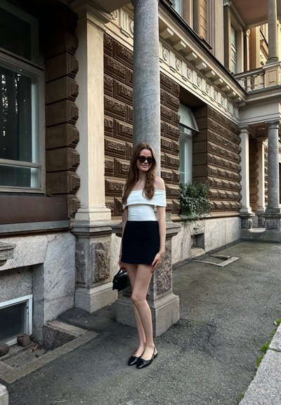 Minifalda negra, blusa blanca sin hombros, gafas de sol negras y zapatos slingback negros. Fondo de mármol y ladrillo ornamentado con detalles de granito.