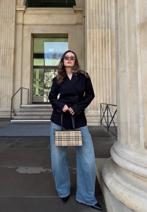 Mujer de pie frente a grandes columnas de piedra, lleva blazer negra, vaqueros de pierna ancha, gafas de sol y sostiene un bolso de cuadros con ambas manos.