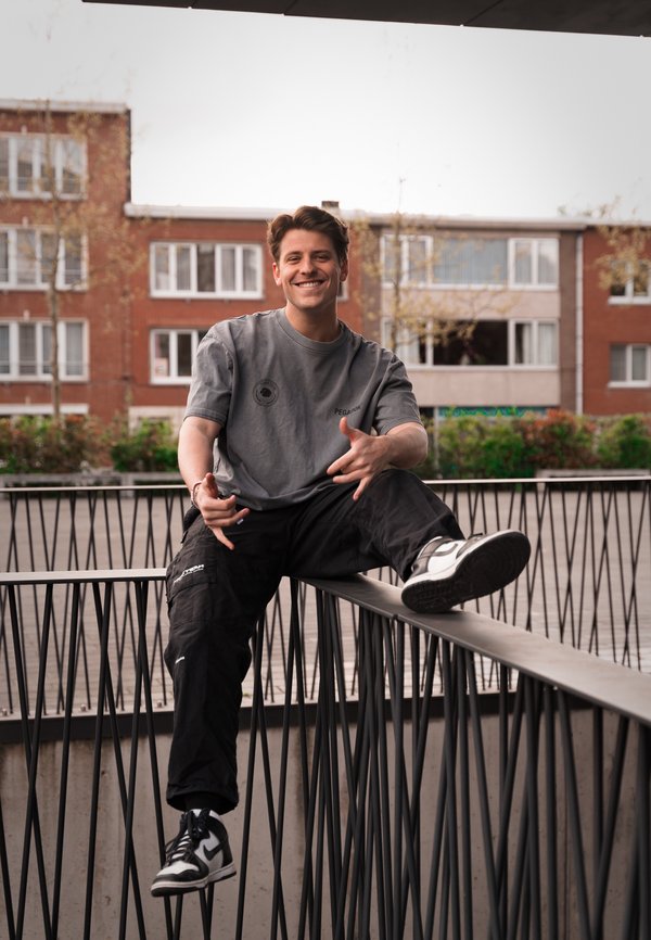 Camiseta gris de manga corta con un logo, pantalones cargo negros con bolsillos laterales y zapatillas deportivas en blanco y negro. Sentado en una barandilla de metal.