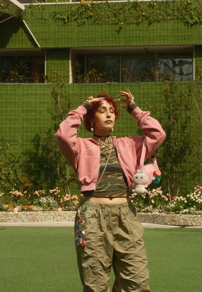 Joven con cabello rojo, chaqueta rosa y camiseta de camuflaje, de pie frente a una pared verde con plantas, ojos cerrados, manos cerca de la cabeza.