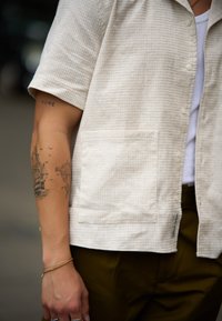 Rövid ujjú, világos bézs ing, texturált mintával, mellzsebbel és gombos záródással. Fehér trikó fölé öltöztetve, zöld nadrággal párosítva.
