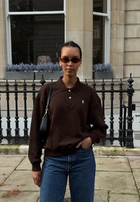 Polo à manches longues marron avec un petit logo, associé à un jean bleu. Les accessoires incluent des lunettes de soleil noires et un petit sac noir.