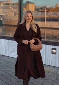 Καφέ μακρύ παλτό πάνω από ένα καφέ ριγέ φόρεμα, κρατώντας μια πλεκτή μπεζ τσάντα. Γυαλιστερές καφέ μπότες μέχρι το γόνατο, σε φόντο από γυαλί και πέτρα.