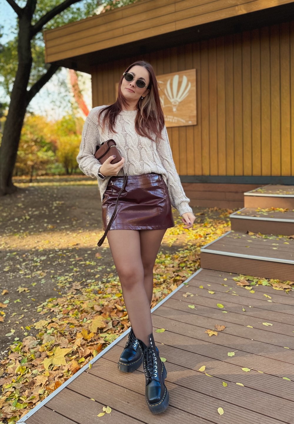 Beige Strickpullover, bordeauxroter Vinyl-Minirock und schwarze Schnürstiefel. Hält eine braune Tasche und steht auf Holzstufen mit Herbstblättern.
