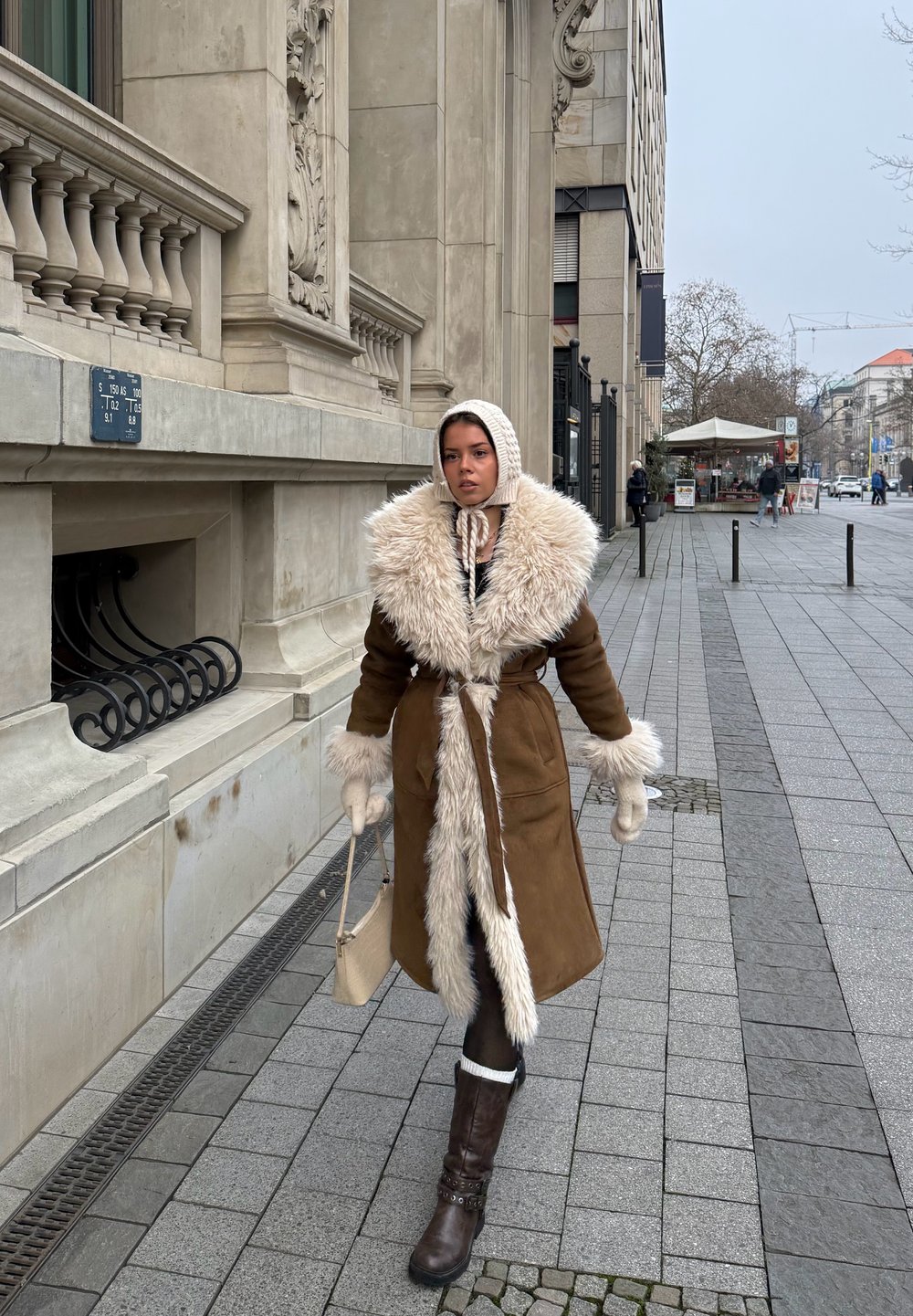 Femme marchant sur un trottoir en ville, portant un manteau marron bordé de fourrure, un bonnet en tricot, des moufles, tenant un sac à main beige, avec un bâtiment historique en arrière-plan.