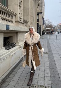 Femme marchant sur un trottoir en ville, portant un manteau marron bordé de fourrure, un bonnet en tricot, des moufles, tenant un sac à main beige, avec un bâtiment historique en arrière-plan.