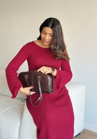 Bruine leren handtas met dubbele handvatten, ritssluiting en een gladde textuur. Model draagt een geribbelde, rode jurk met lange mouwen, zittend op een lichtkleurige bank.