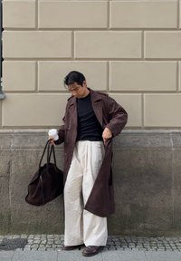 En person i en brun lång kappa och krämfärgade vida byxor står på en trottoar och håller en stor brun väska och en kaffekopp mot en beige vägg.