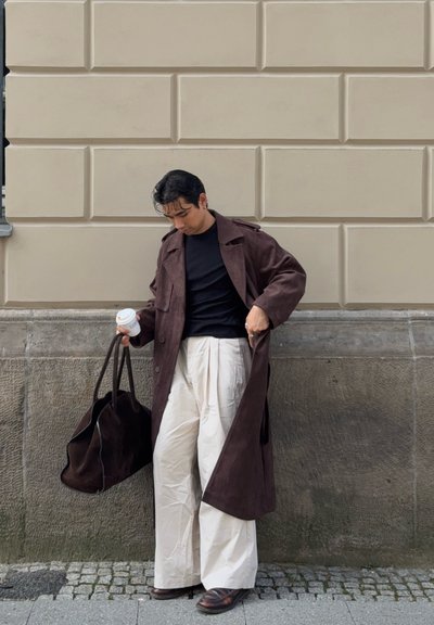 Una persona con un abrigo largo marrón y pantalones anchos crema está de pie en una acera sosteniendo una bolsa grande marrón y una taza de café contra una pared beige.
