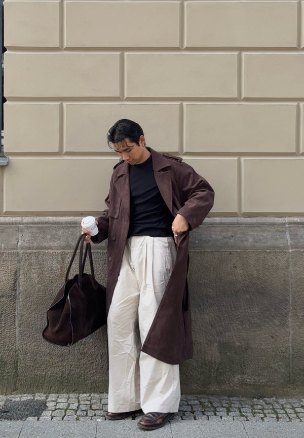 Henkilö, jolla on päällään ruskea pitkä takki ja vaaleanbeige leveät housut, seisoo jalkakäytävällä, pitäen suurta ruskeaa laukkua ja kahvikuppia vaaleanbeigeä seinää vasten.