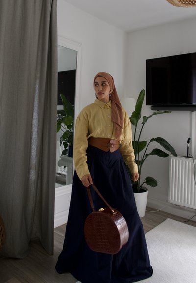 Camisa amarilla de manga larga con cuello, falda azul oscura, cinturón de cuero marrón y bolsa redonda marrón con patrón de cocodrilo. Entorno interior con plantas.