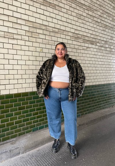 Chaqueta de piel sintética de estampado de leopardo, top corto blanco, jeans anchos azules, botas de plataforma negras, de pie contra una pared de ladrillos texturizada en blanco y verde.