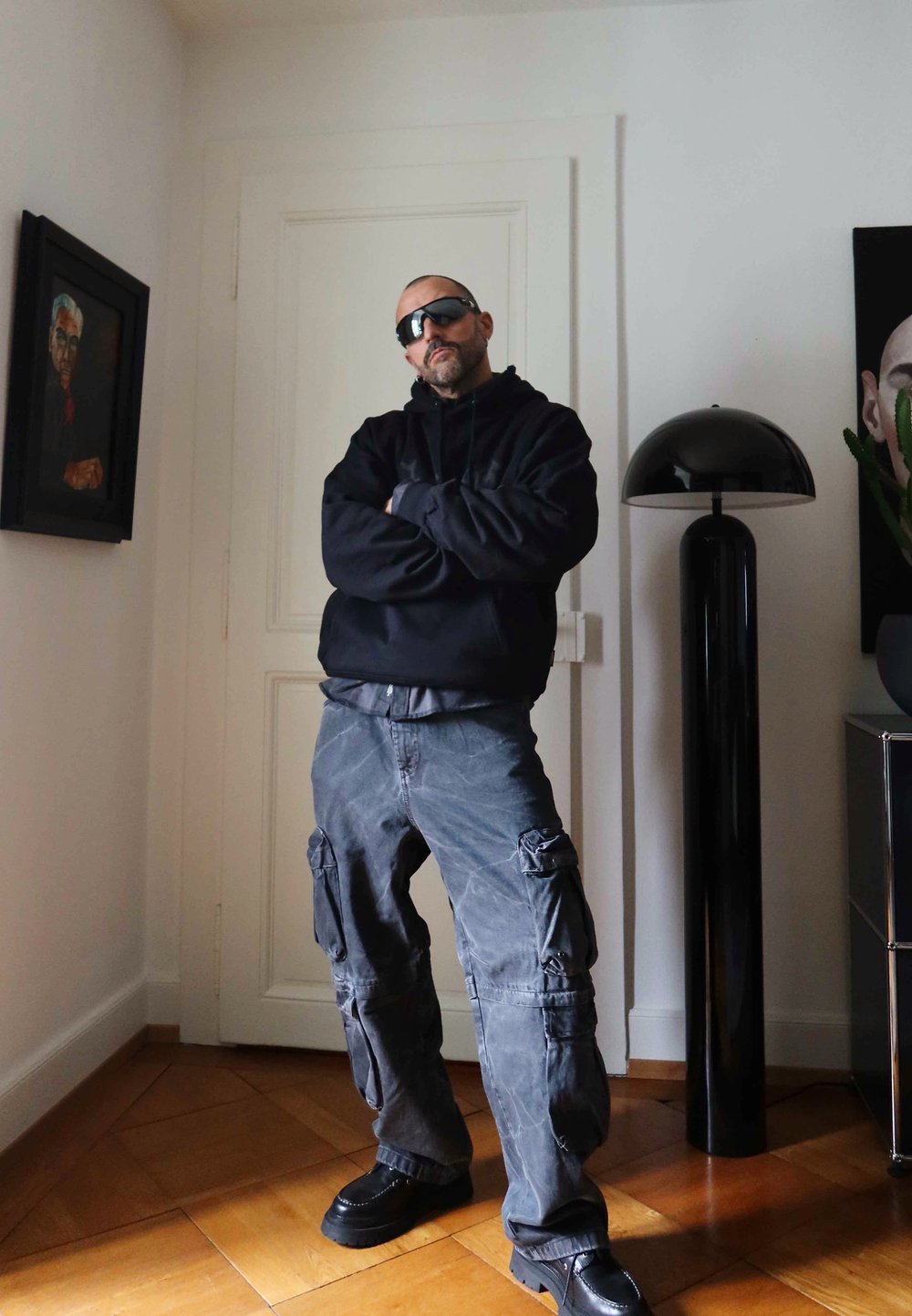 Felpa nera con cappuccio, pantaloni cargo grigi e scarpe nere. In piedi in una stanza con una moderna lampada nera, pavimento in legno e opere d'arte incorniciate sulla parete.