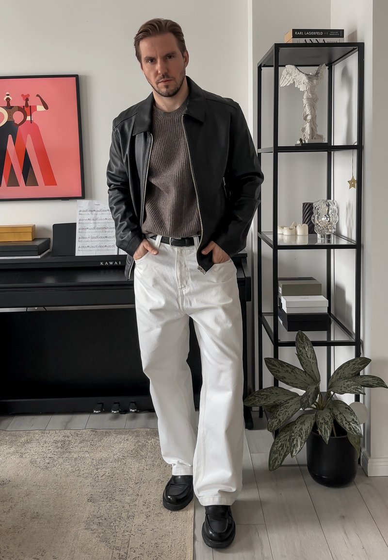 Black leather jacket over a grey ribbed jumper, with white wide-leg trousers and black platform shoes. Standing against a modern interior.